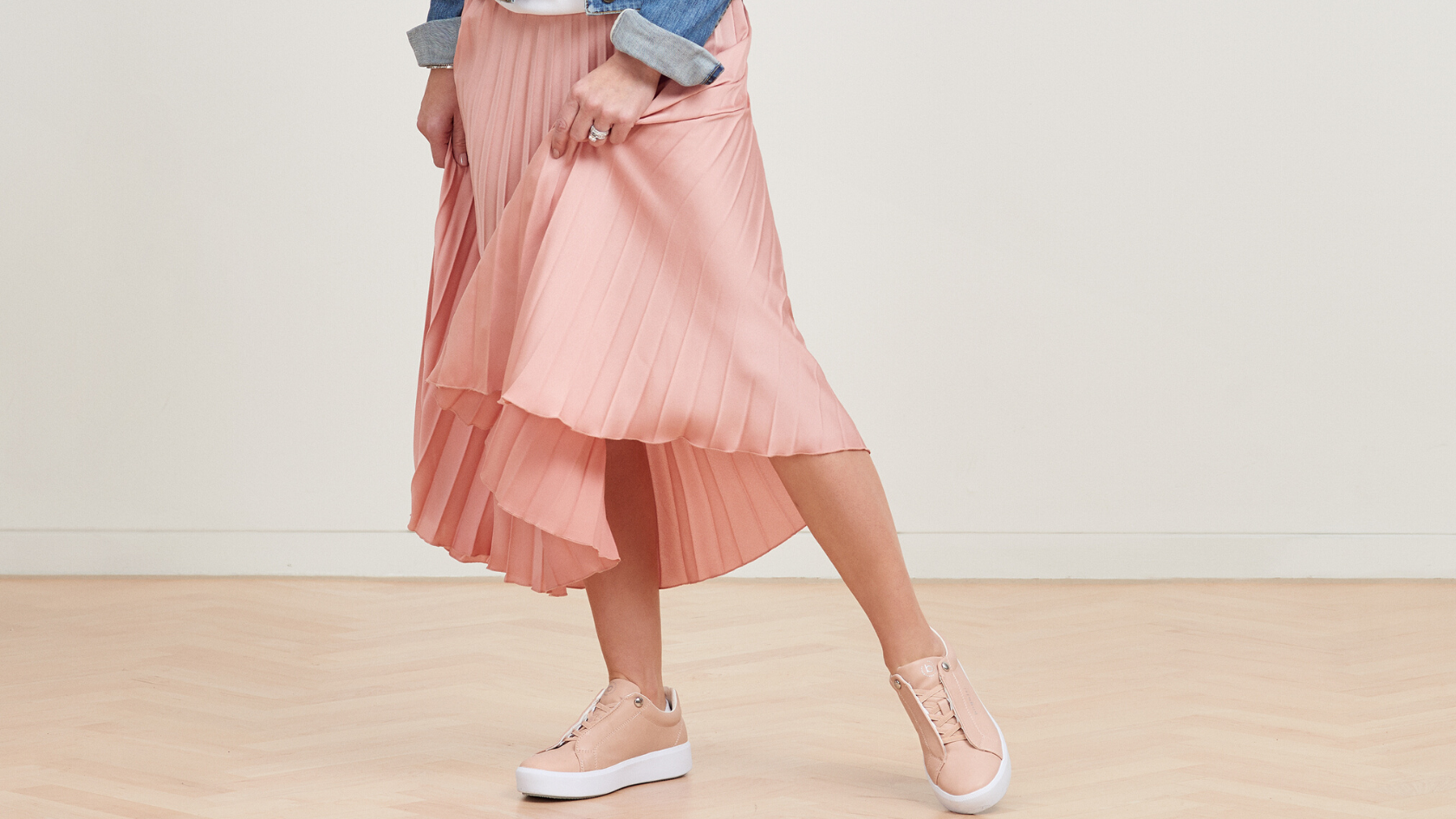 Lady posing in her pink rippled skirt with her pink bugatti sneakers in front of a neutral background