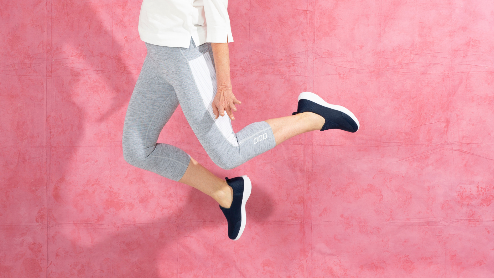 Snap shot of a woman jumping up in front of a pink background wearing slip on sneakers.